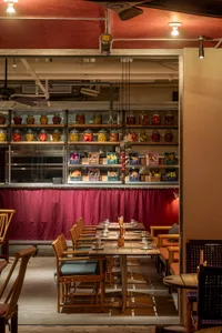 Industrial-style restaurant bar area with wooden tables, metal chairs, glass jars on shelves, and fabric curtains.