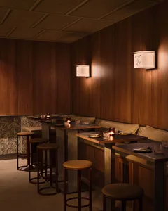 A modern restaurant bar with wooden tables, stools, and booth seating, illuminated by soft lighting.