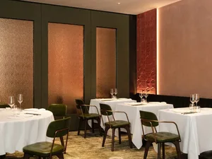 A modern restaurant interior with metal-textured walls, white cloth dining tables, and green chairs.