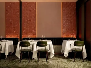 A well-lit, modern restaurant dining room with textured brick walls, green velvet chairs, and white tablecloths.