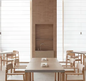 A modern restaurant interior with minimalist design, featuring wooden tables, stone walls, and large windows.