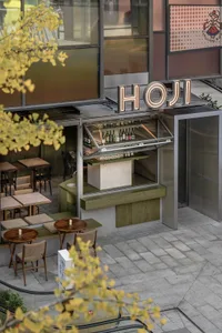 Outdoor seating area of a modern restaurant with metal and wood tables and chairs, glass window display, and neon sign.