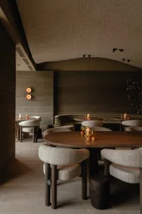 A minimalist restaurant interior featuring wooden tables and chairs, fabric seating, concrete walls, and metal elements.