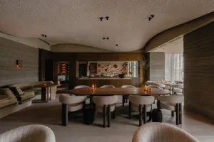Modern restaurant interior with wooden tables, fabric upholstered chairs, and a bar area with a metal counter.