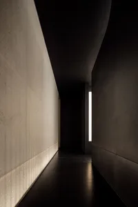 A minimalist corridor with concrete walls and floor, featuring a single illuminated window at the far end.