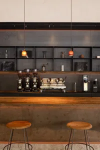 A modern coffee shop interior with a wooden counter, concrete wall, black metal shelving, and coffee-bar setup.