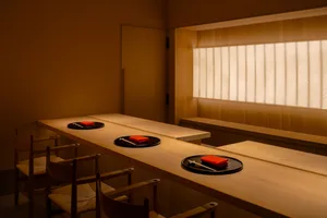 A wooden restaurant counter with black plates, red napkins, and wooden chairs under a window with paper blinds.