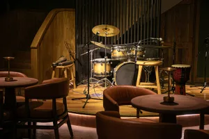 A live music venue featuring a drum set on a stage with wooden flooring and acoustic panels.