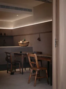 A minimalist residential kitchen with wood and concrete materials, featuring a basket of fruits and a vintage table and chair