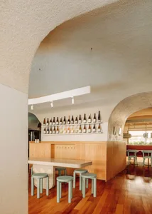 Light wood restaurant bar counter with green stools and bottles lined up on shelves against a textured white wall.
