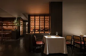 An elegant, dimly lit dining room with a well-lit wine collection in the background and a single table set for a meal.