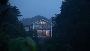 A minimalist house with a glass and wood exterior, surrounded by trees at dusk.