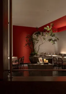 A modern restaurant interior with red walls and a potted plant, tables set with white tablecloth, candles, and glasses.