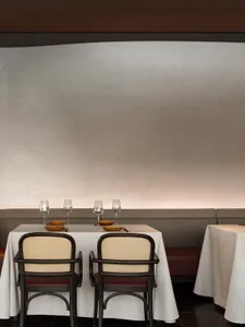 A modern restaurant dining room with white tablecloths, wooden chairs, and minimalist decor.