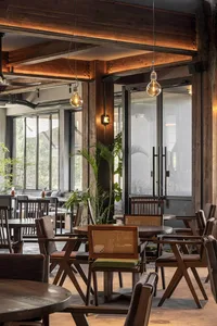 Restaurant interior with wooden tables and chairs, industrial lighting, and greenery.