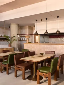 A modern restaurant interior featuring tables and chairs with wooden frames and green leather seats, set against a backdrop o