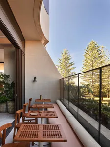 A modern restaurant balcony featuring wood furniture, concrete walls, and glass railings overlooking greenery.