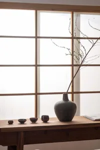 A minimalist vase design placed on a wooden table in front of a Japanese-style shoji window, featuring a natural branch in th
