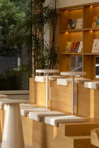 A minimalist cafe interior featuring wooden furniture, potted plants, and a bookshelf filled with books.