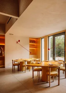A modern, minimalist restaurant interior with wooden tables, yellow chairs, and red accents.