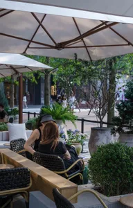 People sitting at tables in a street view cafeteria with wooden tables, metal chairs, concrete planters, and fabric umbrellas