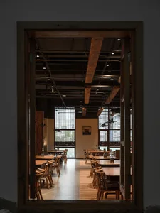 Industrial-style restaurant interior with wooden tables and chairs, large windows, and exposed beams.