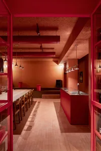 A modern restaurant interior with red cork walls, pendant lights, and a long wooden table with chairs.