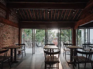 Industrial-style cafe interior featuring wooden tables and chairs, brick walls, and large windows overlooking greenery.