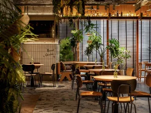 Industrially styled restaurant interior featuring wooden tables and chairs surrounded by green plants and natural light.
