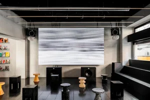 Modern music studio with concrete flooring, stools, and large screen displaying abstract artwork.