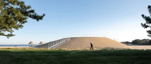 A person walking on a grassy hillside near a sandy hill with a fence on top.