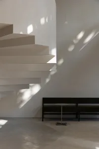 A modern staircase with concrete steps and shadows cast on a white wall, illuminated by natural light from the side.