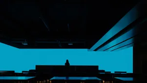 A modern restaurant interior with dark steel tables and seating, illuminated by a large blue screen.