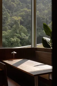 A minimalist wooden table with a lamp, set against a large window overlooking lush greenery.