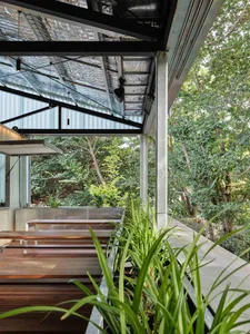 Modern patio featuring a metallic roof structure, potted plants, and wooden benches, surrounded by lush greenery.