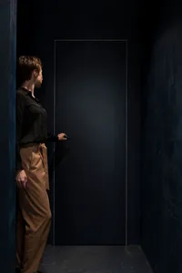 A minimalist photograph of a person holding a black door handle in a dark, textured hallway.