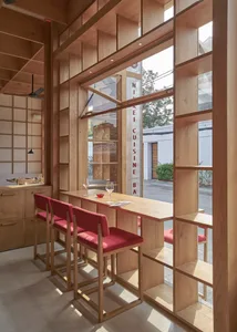 A modern restaurant interior featuring a wooden bar and seating area with red cushioned stools, large windows, and a minimali