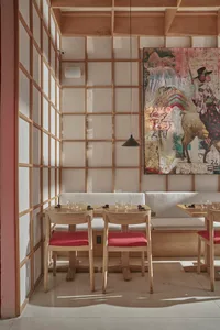 Modern restaurant interior with wooden tables and red cushions, featuring a colorful artwork on the wall.