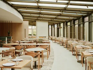 A modern restaurant interior with wooden tables, chairs, and glass panels, featuring a spacious and airy atmosphere.