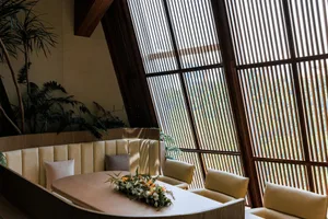 A modern restaurant interior featuring wooden furniture and large windows with slatted shades.