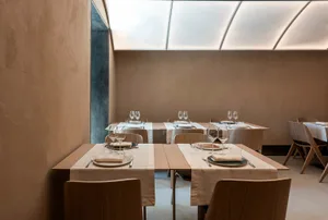 Well-lit minimalist restaurant interior with wooden tables, chairs, and white tablecloths.