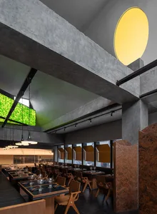 An industrial-style restaurant interior with concrete walls, wooden furniture, and a large green screen.