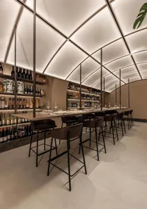 A modern restaurant bar with sleek design elements, including a long table with seating, wine bottles on shelves, and a conte