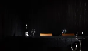 A minimalist restaurant interior with dark wood walls and tables, featuring a single bottle of sake and wine glasses, creatin