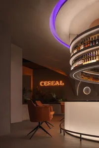 A modern restaurant bar with concrete, glass, and wood finishes, featuring a circular lighting fixture and a neon sign.