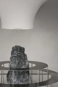 A black rock-like object encased in a circular metal frame, illuminated by a large, white ceiling light in a minimalist indus