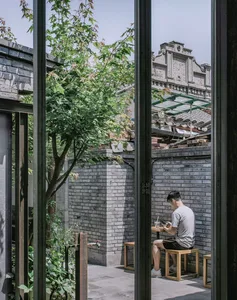 A man sits at a table in a courtyard made of bricks and surrounded by wooden furniture and greenery.