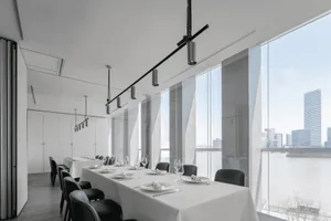 A minimalist restaurant interior with white walls and floors, featuring tables set with white tablecloths and black chairs.