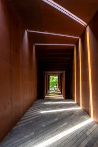 A narrow hallway lined with industrial-style wood flooring and metallic walls, leading to an open area with greenery.
