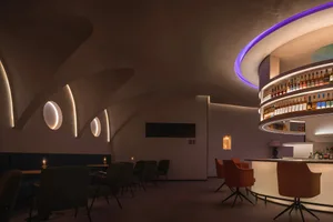A modern industrial restaurant bar with concrete walls and ceiling, featuring a curved bar area with illuminated shelves and 
